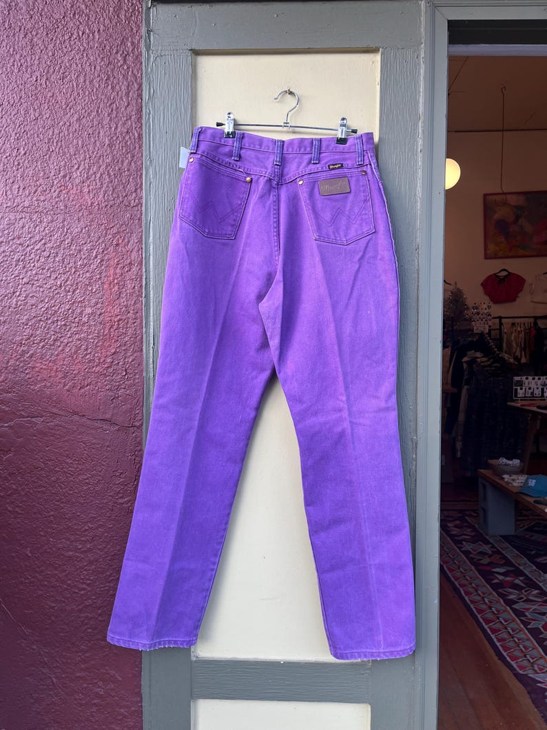 May include: Bright purple Wrangler jeans hanging on a metal hanger. The jeans have a classic five-pocket design with a brown leather patch on the back pocket. The jeans are displayed in a doorway with a purple wall on the left.