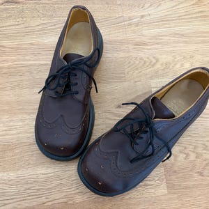 Può includere: Un paio di scarpe Oxford in pelle marrone con lacci neri. Le scarpe presentano decorazioni traforate e una suola spessa e scura. Sono appoggiate su una superficie di legno chiaro.