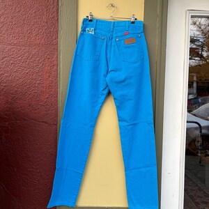 May include: Bright blue denim jeans hanging on a metal hanger. The jeans have a classic five-pocket design with a leather patch and a small embroidered detail on the back pocket. The jeans are displayed against a yellow door frame and a red wall.