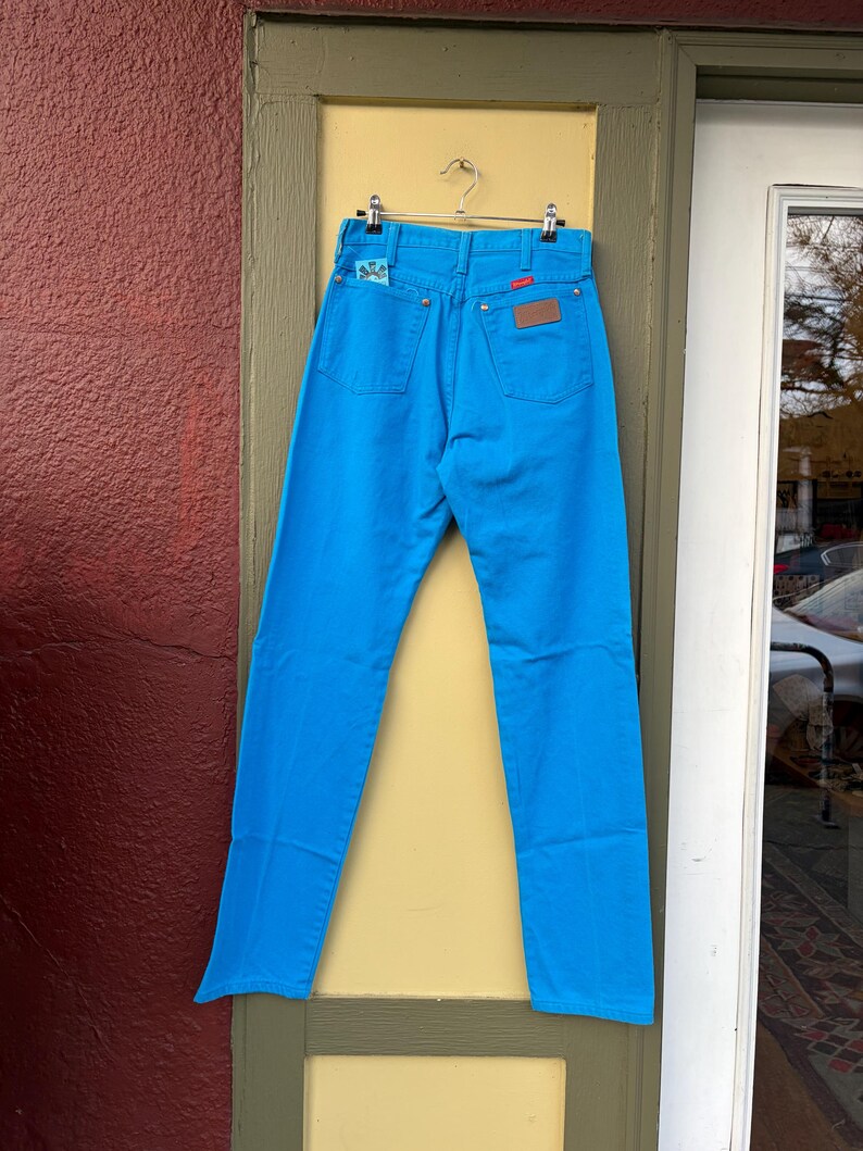 May include: Bright blue denim jeans hanging on a metal hanger. The jeans have a classic five-pocket design with a leather patch and a small embroidered detail on the back pocket. The jeans are displayed against a yellow door frame and a red wall.