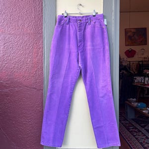 May include: A pair of vibrant purple denim pants hangs on a silver hanger. The high-waisted jeans have a classic five-pocket design and a straight leg. The pants are displayed in an open doorway against a neutral background.
