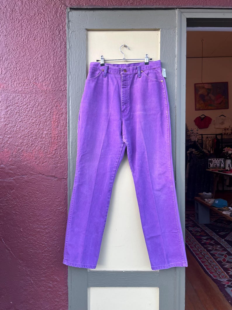 May include: A pair of vibrant purple denim pants hangs on a silver hanger. The high-waisted jeans have a classic five-pocket design and a straight leg. The pants are displayed in an open doorway against a neutral background.