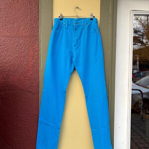 May include: Bright blue denim jeans hanging on a metal hanger. The jeans have a classic five-pocket design with a button and zipper closure. The jeans are displayed against a yellow and green door frame.