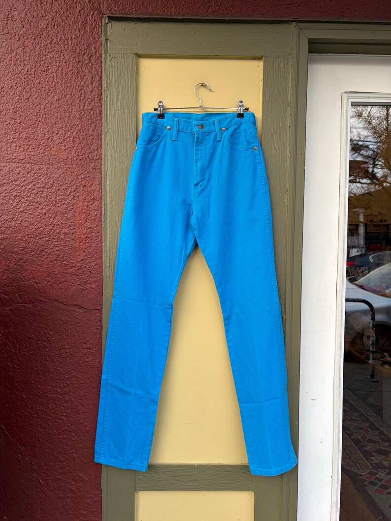 May include: Bright blue denim jeans hanging on a metal hanger. The jeans have a classic five-pocket design with a button and zipper closure. The jeans are displayed against a yellow and green door frame.