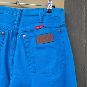 May include: Close-up of bright blue Wrangler jeans hanging on a metal hanger. The jeans have a brown leather patch with the Wrangler logo on the back pocket and a red Wrangler tag. The jeans are made of a sturdy denim material.