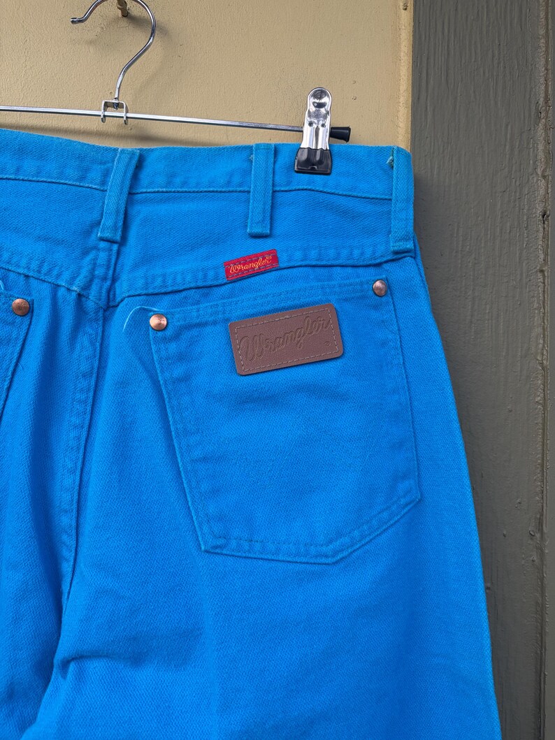 May include: Close-up of bright blue Wrangler jeans hanging on a metal hanger. The jeans have a brown leather patch with the Wrangler logo on the back pocket and a red Wrangler tag. The jeans are made of a sturdy denim material.