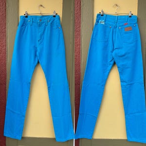 May include: Bright turquoise denim jeans, front and back views, hanging on a metal hanger. The jeans have a classic five-pocket design, a button and zipper closure, and a brown leather patch on the back. The jeans are a vibrant, solid color.