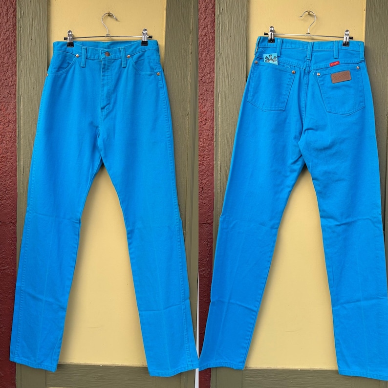 May include: Bright turquoise denim jeans, front and back views, hanging on a metal hanger. The jeans have a classic five-pocket design, a button and zipper closure, and a brown leather patch on the back. The jeans are a vibrant, solid color.