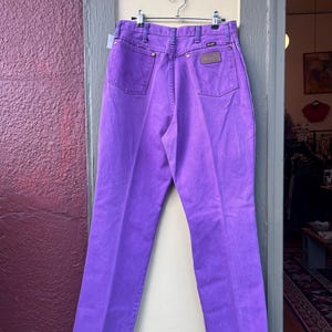 May include: Bright purple Wrangler jeans hanging on a metal hanger. The jeans have a classic five-pocket design with a brown leather patch on the back. The stitching is a darker purple, and the jeans are made of a sturdy denim material.