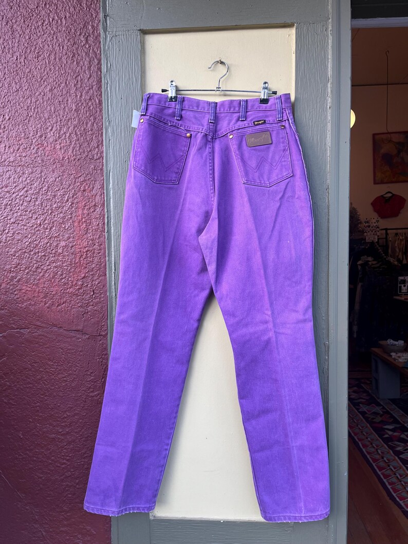 May include: Bright purple Wrangler jeans hanging on a metal hanger. The jeans have a classic five-pocket design with a brown leather patch on the back. The stitching is a darker purple, and the jeans are made of a sturdy denim material.
