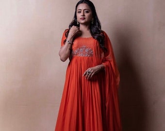May include: A woman wearing a long, orange dress with a silver embroidered neckline and a matching sheer scarf. The dress has a fitted bodice and a flowing skirt.