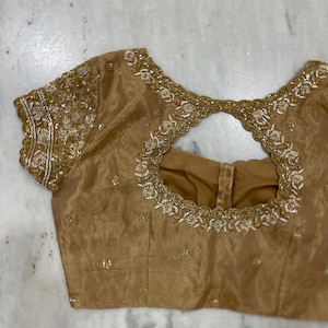Puede incluir: Una blusa dorada con intrincados bordados alrededor del escote y los puños de las mangas. La blusa presenta un diseño de ojo de cerradura en la parte delantera y delicados adornos. La tela tiene un brillo sutil.