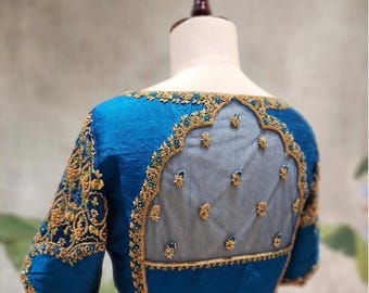 May include: A teal-blue blouse with intricate gold embroidery on the sleeves and neckline. The back features a sheer panel with delicate floral embellishments. The blouse is displayed on a mannequin.