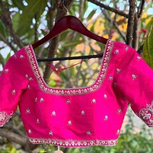 May include: A vibrant fuchsia blouse with short sleeves, adorned with intricate embroidery along the neckline and sleeve cuffs. The embroidery features delicate floral and bird motifs, adding a touch of elegance to the garment. The blouse is hanging on a wooden hanger.
