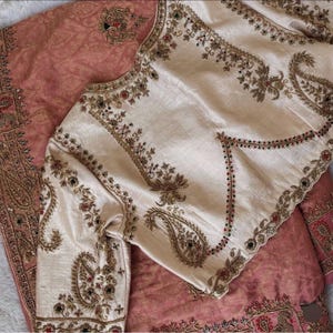 May include: A cream-coloured blouse featuring intricate gold embroidery and small red and green bead accents, layered over a dusty rose fabric with a subtle tonal pattern. The blouse has long sleeves and a structured neckline with detailed floral motifs.