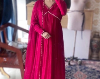 Puede incluir: Un vestido largo de color rosa con cuello en V y mangas largas. El vestido tiene un diseño bordado blanco alrededor del cuello y las mangas. El vestido está hecho de una tela suave y fluida.