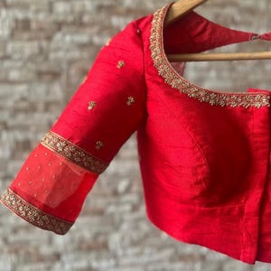 May include: A red silk blouse with gold embroidery and a sheer overlay on the sleeves. The blouse has a round neckline and a fitted bodice.