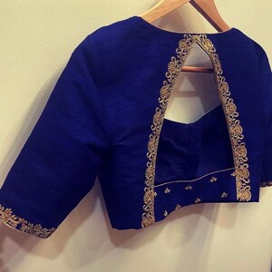 Op de afbeelding: Een blauwe blouse met gouden borduurwerk rond de halslijn en mouwen. De blouse heeft een sleutelgat opening in de rug.