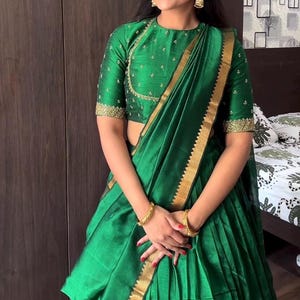 May include: A green sari with gold edging and a matching green blouse. The blouse features short sleeves and gold embroidery around the neckline and sleeves. The sari is draped, with the pleats visible.