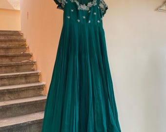 May include: A teal green maxi dress with white embroidery. The dress has a round neckline and short sleeves. The dress is pleated and has a flowy silhouette.