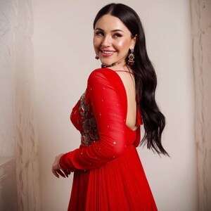 May include: A woman in a red dress with long sleeves and a low back. The dress is pleated and has a beaded design on the bodice. She is wearing earrings and a necklace.