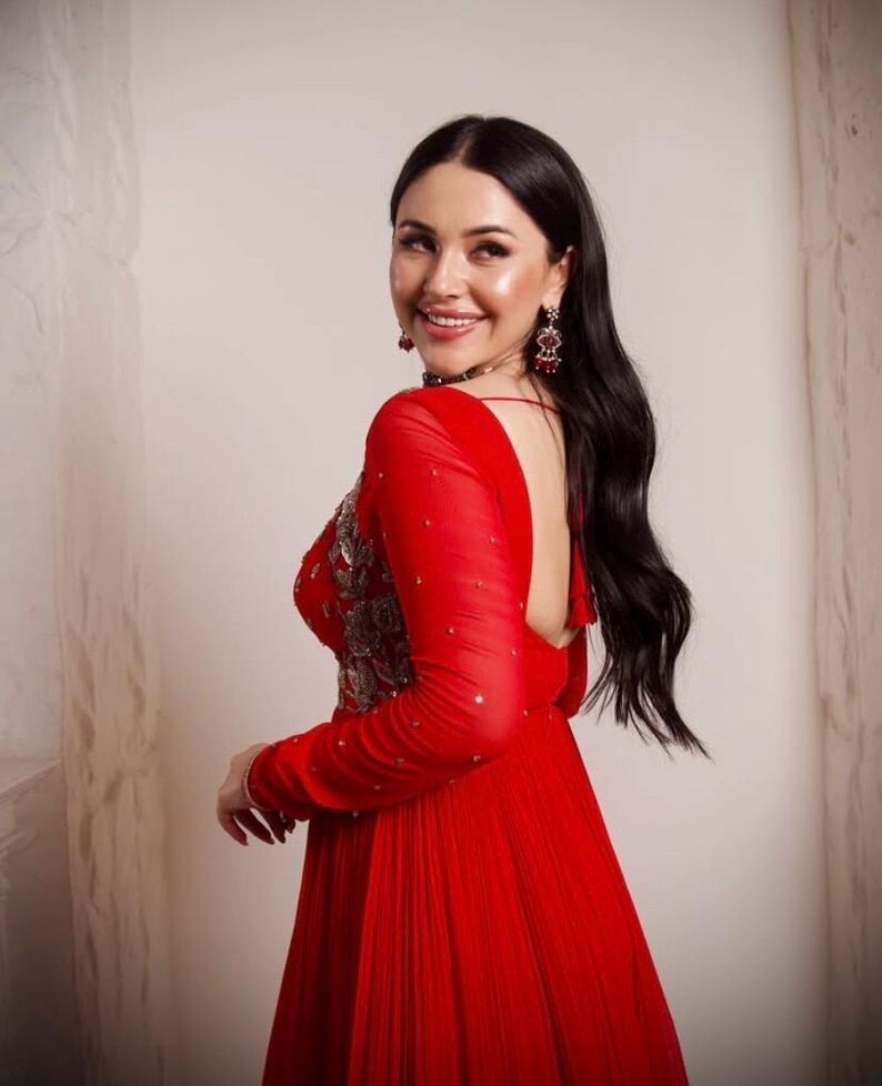 May include: A woman in a red dress with long sleeves and a low back. The dress is pleated and has a beaded design on the bodice. She is wearing earrings and a necklace.