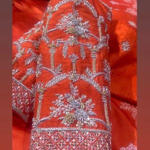 K&ouml;nnte beinhalten: Nahaufnahme eines leuchtend orangefarbenen &Auml;rmels, aufwendig mit Silber- und Goldf&auml;den bestickt. Das Design zeigt florale und geometrische Muster, die eine luxuri&ouml;se und detaillierte &Auml;sthetik schaffen. Der &Auml;rmel hat eine detaillierte Bord&uuml;re.