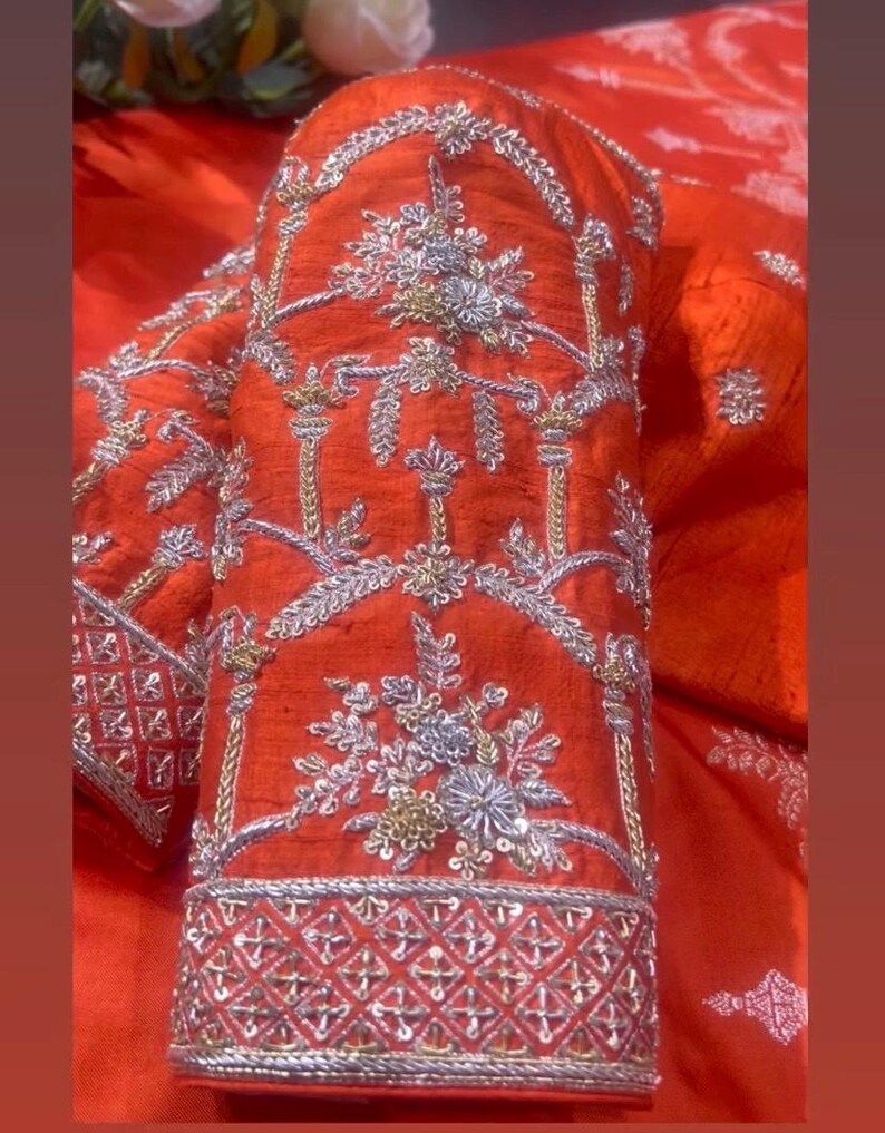 K&ouml;nnte beinhalten: Nahaufnahme eines leuchtend orangefarbenen &Auml;rmels, aufwendig mit Silber- und Goldf&auml;den bestickt. Das Design zeigt florale und geometrische Muster, die eine luxuri&ouml;se und detaillierte &Auml;sthetik schaffen. Der &Auml;rmel hat eine detaillierte Bord&uuml;re.