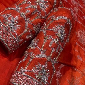 K&ouml;nnte beinhalten: Nahaufnahme von zwei roten Stoff&auml;rmeln mit aufwendiger Silber- und Goldstickerei. Die &Auml;rmel weisen florale und geometrische Muster mit einem detaillierten Bord&uuml;rendesign auf. Der Stoff hat einen leichten Glanz, der die satte Farbe und Textur verst&auml;rkt.