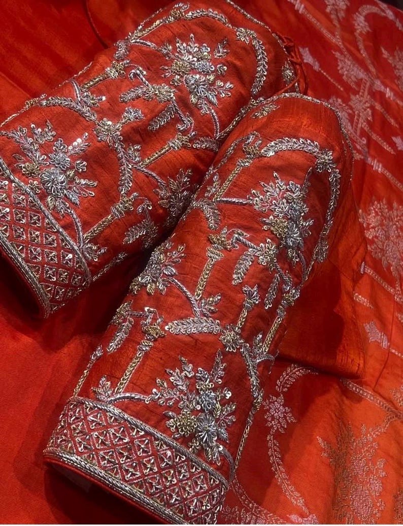 K&ouml;nnte beinhalten: Nahaufnahme von zwei roten Stoff&auml;rmeln mit aufwendiger Silber- und Goldstickerei. Die &Auml;rmel weisen florale und geometrische Muster mit einem detaillierten Bord&uuml;rendesign auf. Der Stoff hat einen leichten Glanz, der die satte Farbe und Textur verst&auml;rkt.