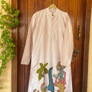 May include: A long-sleeved, light pink kurta with a button-up collar and gold embroidery. The kurta features a hand-painted design of a peacock, a palm tree, and a figure playing a flute.
