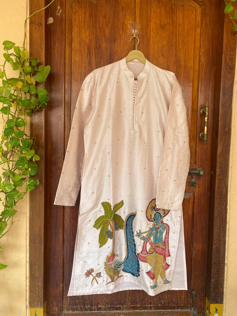 May include: A long-sleeved, light pink kurta with a button-up collar and gold embroidery. The kurta features a hand-painted design of a peacock, a palm tree, and a figure playing a flute.