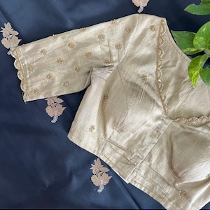 Puede incluir: Una blusa beige bordada con escote festoneado y mangas cortas. La blusa está decorada con pequeñas lentejuelas y cuentas doradas.