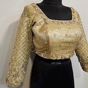 Puede incluir: Una blusa dorada de manga larga con intrincados bordados y abalorios. El escote cuadrado y las mangas están adornados con detalles. La blusa se exhibe en un maniquí negro.