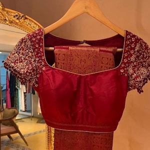 May include: A maroon blouse with intricate gold embroidery on the short sleeves. The square neckline and bodice are a solid maroon color. A matching maroon and gold patterned fabric is visible behind the blouse.