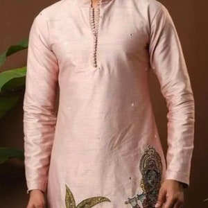 May include: A pink long-sleeved kurta with a traditional Indian design featuring a peacock, a palm tree, and a figure playing a flute. The kurta has a mandarin collar and a button closure.