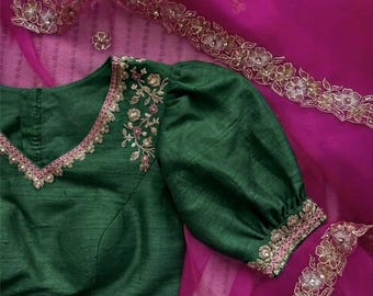 May include: A dark green blouse with a V-neckline and puffed sleeves. The neckline and sleeve cuffs are trimmed with a delicate floral embroidery and pink accents. A sheer pink fabric with floral trim is in the background.
