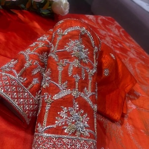 K&ouml;nnte beinhalten: Nahaufnahme eines orangefarbenen Kleidungsst&uuml;cks mit aufwendiger Silber- und Goldstickerei. Das Design zeigt detaillierte Blumenmuster und geometrische Bord&uuml;ren. Der Stoff hat ein gl&auml;nzendes Aussehen und ist luxuri&ouml;s.