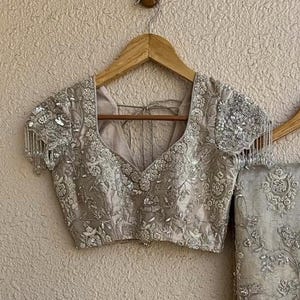 Op de afbeelding: Een zilver-grijze blouse met korte mouwen, gedetailleerde bloemenborduurwerk en kralenfranjes op de mouwen. De blouse heeft een diepe V-hals en wordt getoond op een houten hanger, wat de delicate details benadrukt.