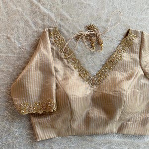 Puede incluir: Blusa beige a rayas con cuello en V. El escote y los puños de las mangas están adornados con bordados dorados y pequeñas cuentas. La blusa tiene mangas tres cuartos y un detalle de cordón.