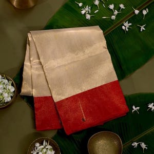 May include: A folded textile piece featuring a large, shimmering champagne-colored section and a wide, solid red border at the bottom. The fabric has a subtle, reflective sheen and a smooth texture, presented as a rectangular, layered garment.