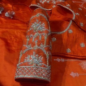K&ouml;nnte beinhalten: Nahaufnahme eines orangefarbenen Seidenkleidungsst&uuml;cks mit aufwendiger silberner Stickerei. Die Stickerei zeigt florale und geometrische Muster. Das Kleidungsst&uuml;ck scheint eine traditionelle indische Bluse oder ein Oberteil zu sein, mit detaillierten &Auml;rmeln und Ausschnitt.