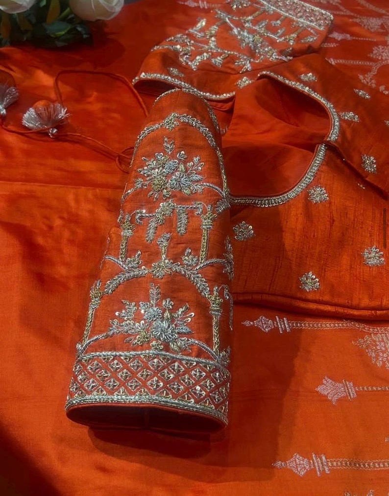 K&ouml;nnte beinhalten: Nahaufnahme eines orangefarbenen Seidenkleidungsst&uuml;cks mit aufwendiger silberner Stickerei. Die Stickerei zeigt florale und geometrische Muster. Das Kleidungsst&uuml;ck scheint eine traditionelle indische Bluse oder ein Oberteil zu sein, mit detaillierten &Auml;rmeln und Ausschnitt.