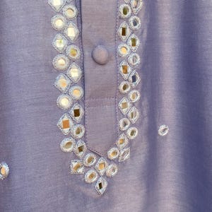 K&ouml;nnte beinhalten: Eine Nahaufnahme eines lavendelfarbenen Stoffes mit silbernen Spiegelverzierungen. Der Stoff hat einen dezenten Glanz und ist mit einem Muster aus kleinen, runden Spiegeln verziert.