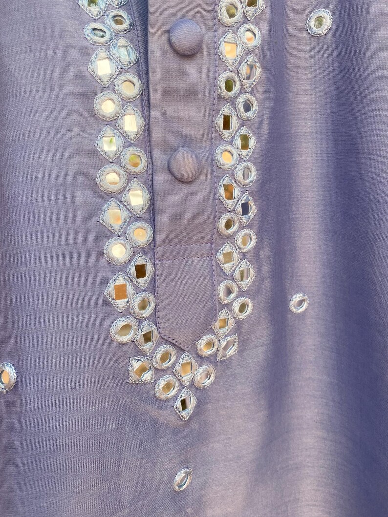 K&ouml;nnte beinhalten: Eine Nahaufnahme eines lavendelfarbenen Stoffes mit silbernen Spiegelverzierungen. Der Stoff hat einen dezenten Glanz und ist mit einem Muster aus kleinen, runden Spiegeln verziert.