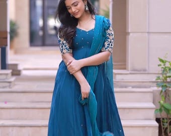 Puede incluir: Una mujer lleva un vestido largo de manga larga azul turquesa con bordados dorados en las mangas y el escote. El vestido tiene una falda fluida y se combina con una bufanda azul turquesa a juego.