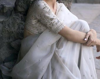 Può includere: Un sari bianco con delicate decorazioni argentate e una blusa abbinata con ricami intricati. Il sari è drappeggiato con eleganza, creando morbide pieghe e una silhouette fluida. La blusa presenta polsini delle maniche dettagliati.