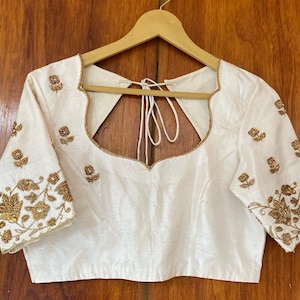 Peut inclure: Chemise en soie blanche avec des broderies dorées représentant des motifs floraux. La chemise a un col rond et des manches courtes.