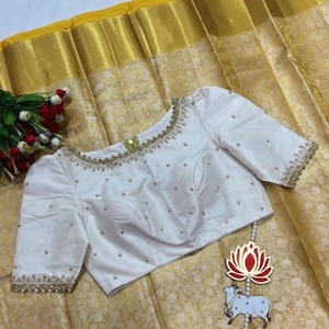 May include: White embroidered blouse with pearl embellishments and a scalloped neckline. The blouse is on a gold and white patterned fabric.