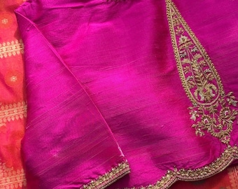 May include: A vibrant magenta blouse featuring intricate gold embroidery along the neckline, sleeve cuffs, and a large teardrop-shaped panel on the front. The garment is crafted from a textured fabric with a subtle sheen and scalloped hem edges.